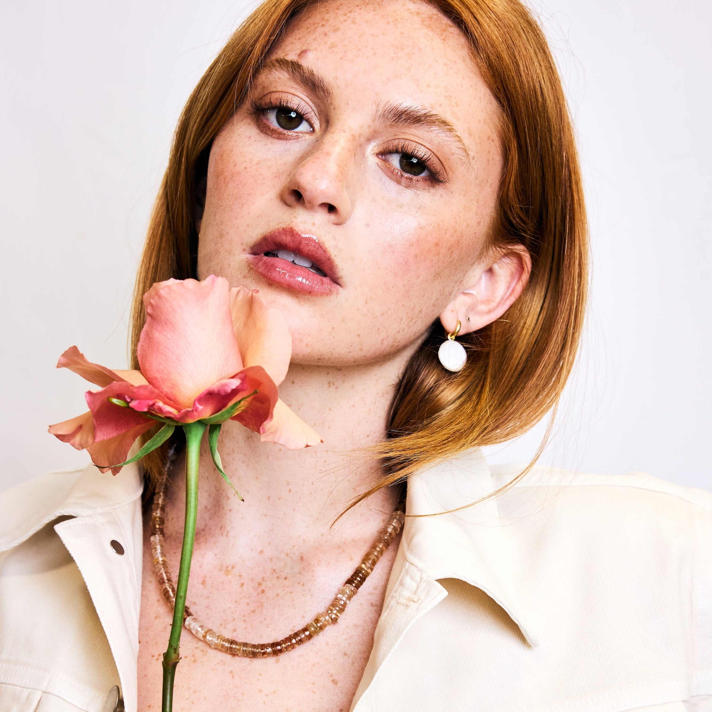A close-up portrait of a woman with red hair wearing a cream jacket and holding a pink rose near her face, set against a plain light background.